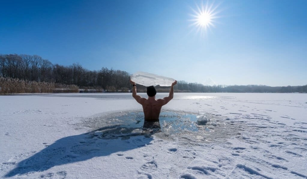 Un hombre dentro de un lago helado, sosteniendo un trozo de hielo sobre su cabeza como parte de una estimulante experiencia de inmersión en agua fría, mostrando la importancia de los baños de hielo para la recuperación y rendimiento