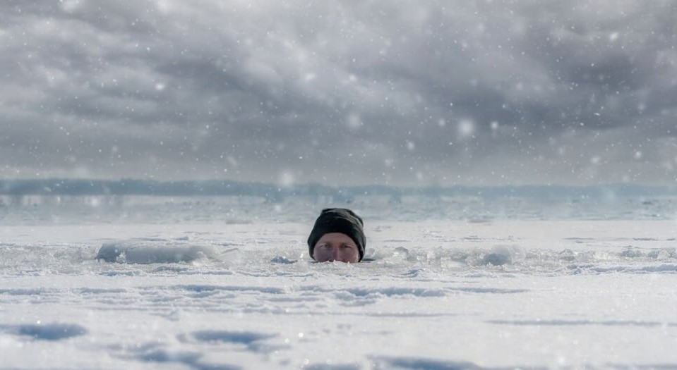 La parte superior de la cabeza de un hombre que emerge de un lago helado, haciendo contacto visual con la cámara, simboliza la importancia de los baños de hielo para la recuperación y rendimiento